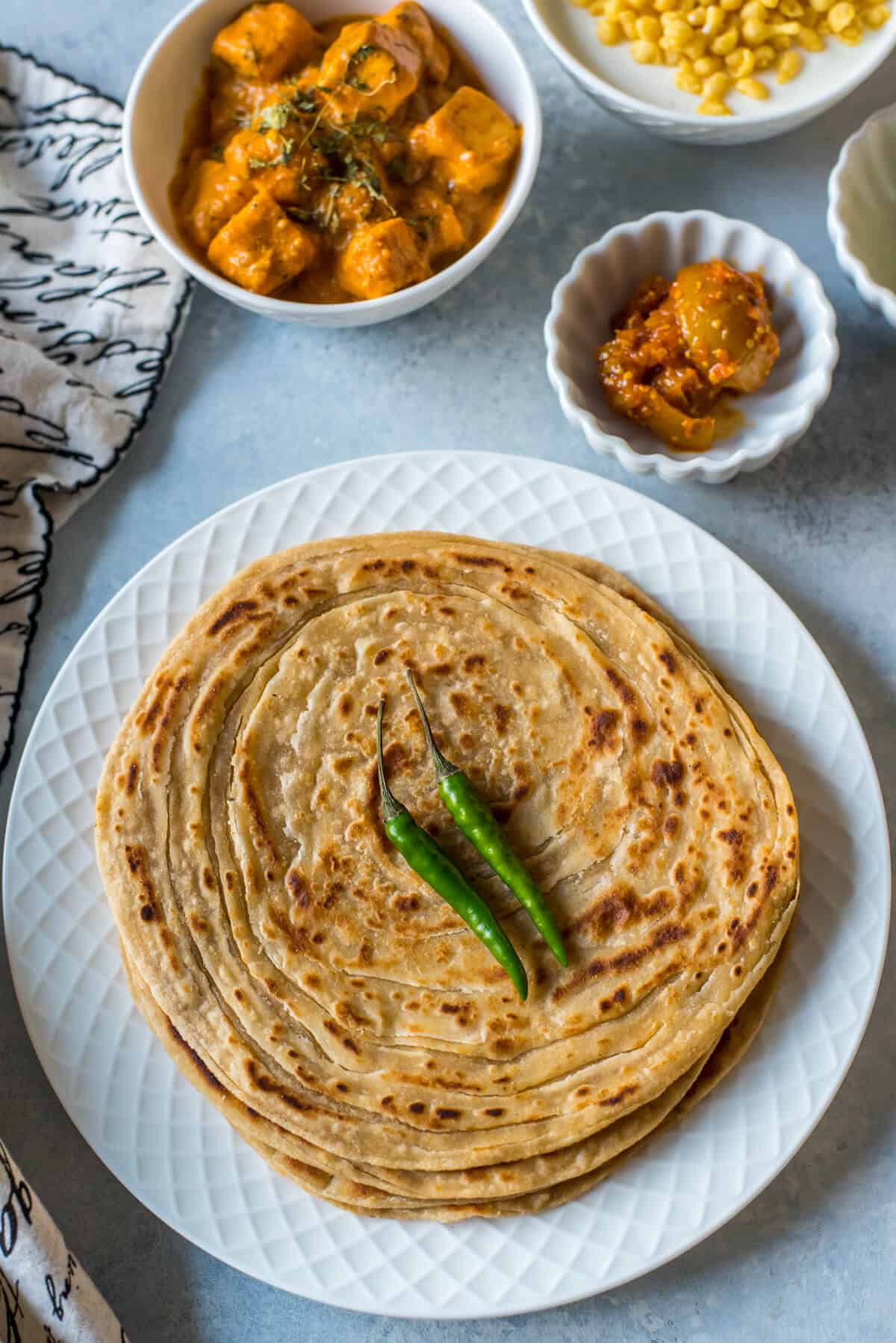 A white plate with a stack of lacha paratha topped with two green chilies in the front and a few white bowls in the back right corner.