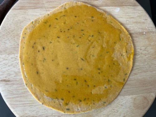 A round wooden cutting board with the rolled out paratha brushed with ghee.