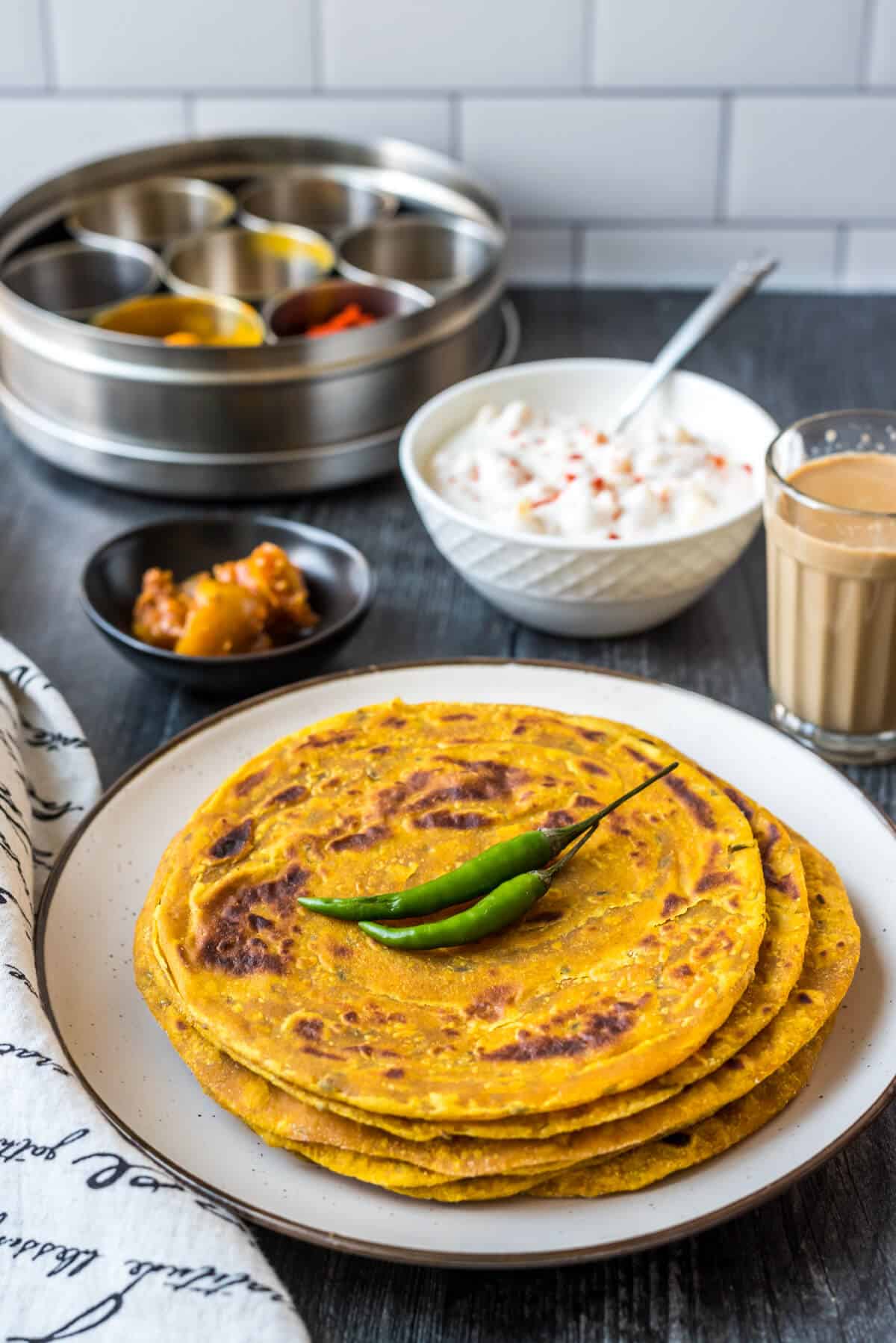 A white plate with a small stack of masala paratha in the front and a cup of team and bowl of raita in the back.