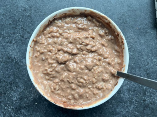 A white mixing bowl with a silver spoon to the right with peanut butter chocolate oatmeal after mixing in the cocoa powder.