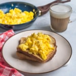 A white plate with a brown rim and a slice of toast topped with fresh scrambled eggs with a skillet in the back left and a glass of tea in the top right.