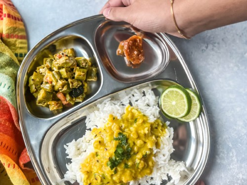 Hands holding a stainless steel tray with dal, rice, and vegetable sides.