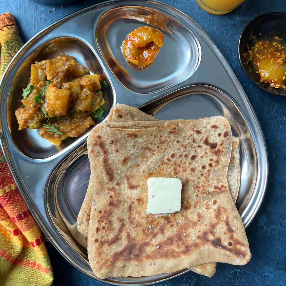 Ajwain paratha sitting on a silver food tray, on a blue counter top. The tray has dividers to separate the curry and mango chutney which is also on the tray. There is a chunk of butter on top of the paratha