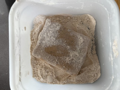 A square flat piece of dough is inside the tub containing flour and has been coated.