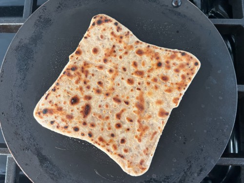 The carom seed paratha is frying in a pan. It has multiple dark spots over it.