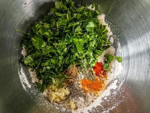 Adding fenugreek leaves and other ingredients to a stainless bowl.