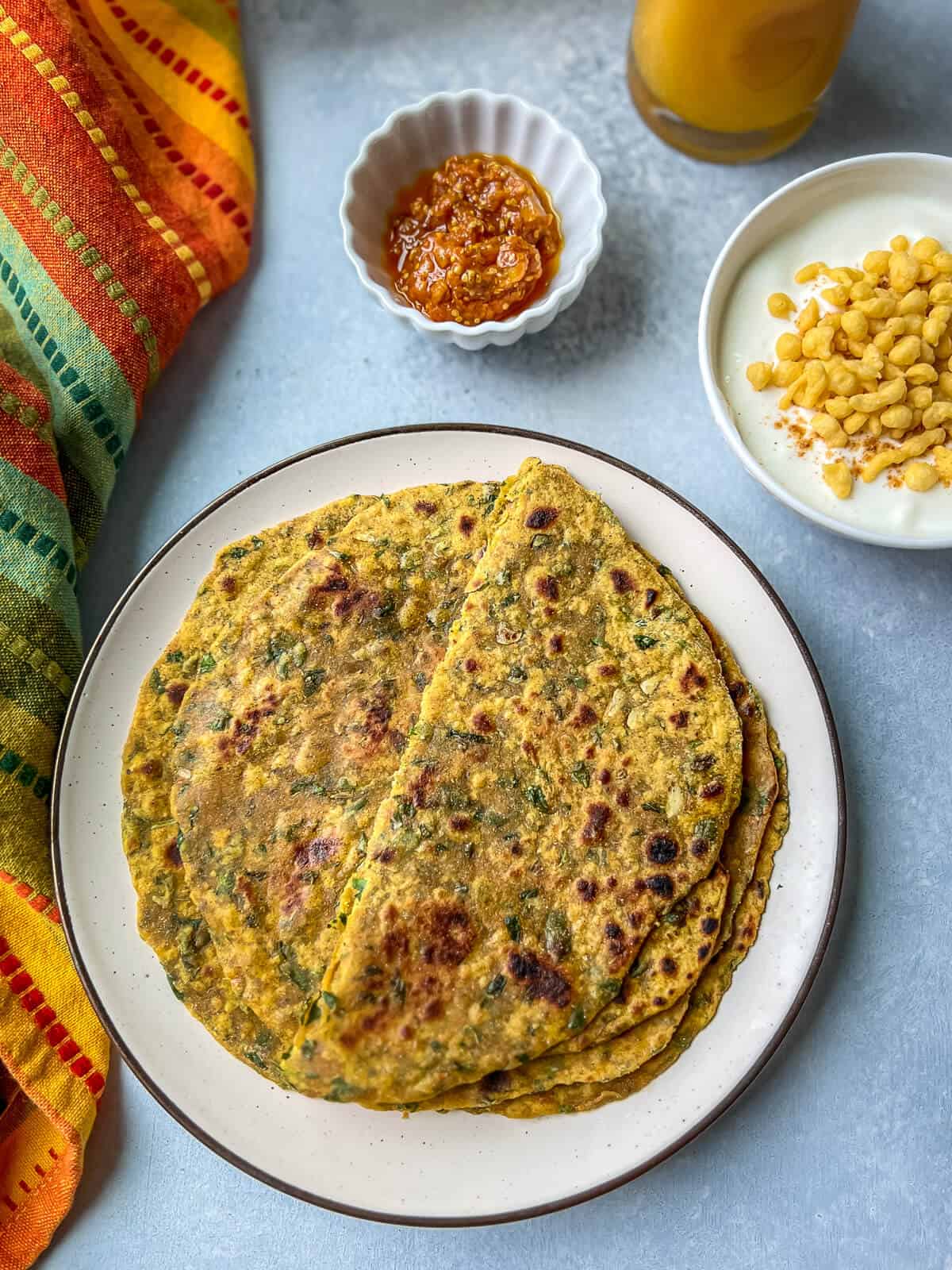 A stack of paratha on a plate, with bowls of raita and pickle on the side.