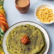 Top down view of layers of spinach paratha on a white plate. Bowl of boodi raita and mug of coffee above.