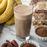A coffee smoothie in a glass with ingredients such as banana, dates, walnuts, coffee and cocoa powder on the side
