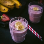 Two glasses of strawberry banana smoothie with sliced bananas on top. With bananas and red and white straws on the side.