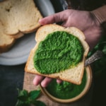 Bread slathered with green chutney is placed on a palm. There are additional bread slices kept on a grey plate.